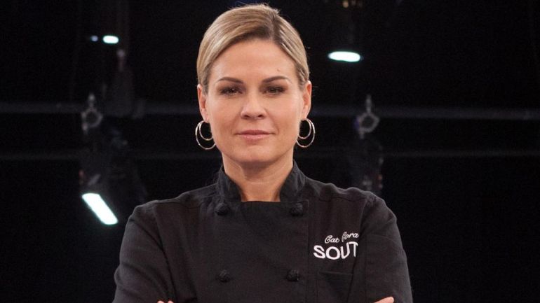 Headshot of Cat Cora, one of the most famous chefs on television, wearing a black chefs uniform and standing in front of a black background.