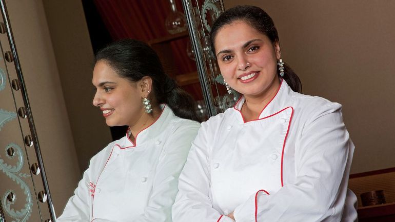 Photo of Maneet Chauhan, one of the most famous chefs, wearing a white uniform and standing in front of a mirror facing the camera.