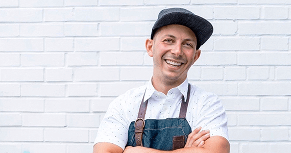 Headshot photo of Matt Basile, one of the most famous chefs on television, standing in front of a white brick wall.