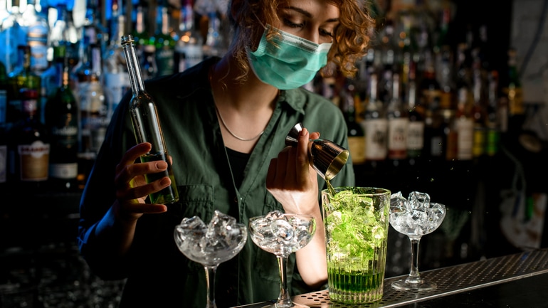 Young bartender in a face mask pouring liquor from beaker into glass with ice