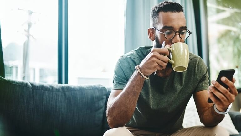 Man sitting on a couch, while drinking coffee, and using a smartphone.