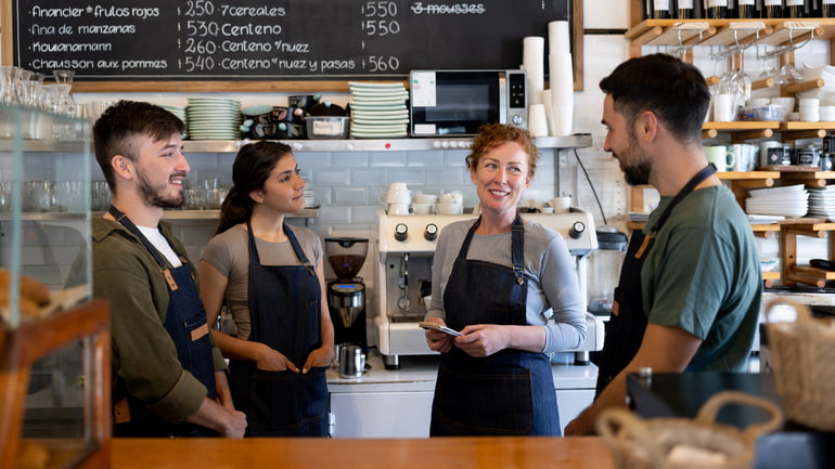A group of two men and two women who work at a cafe are standing behind the counter talking.