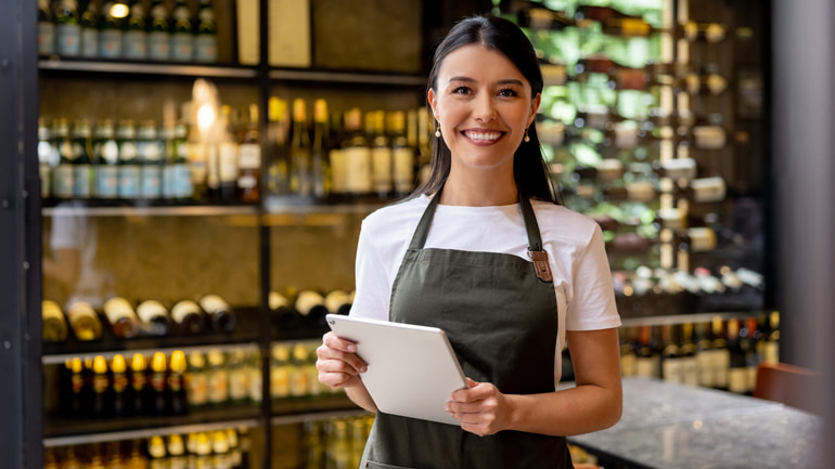Woman wearing a white t-shirt and dark olive colored apron, smiling while holding a tablet with alcohol behind her.