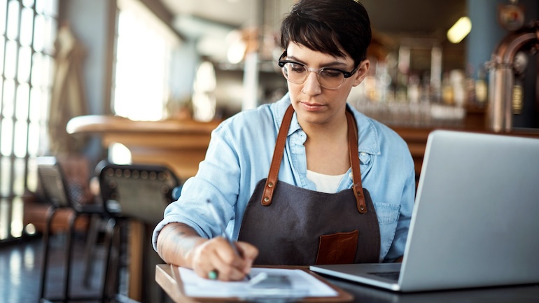 Restaurant staff member writing on a clipboard with a laptop in front of them.