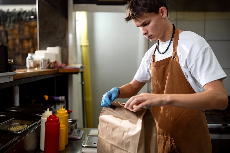 A male teenager packs an online order into a brown paper bag in a fast food kitchen.