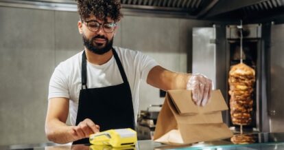Man with curly hair and glasses organizes a food order at the counter using a yellow payments device in a modern restaurant kitchen with food preparation area.