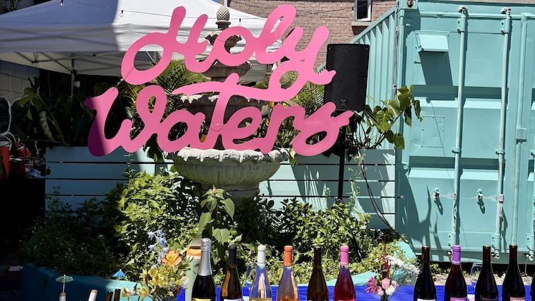 Pink signage at Paradise Grapevine event saying "Holy Waters" behind a table of wine bottles.